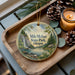 A wooden tray with a decorative ornament featuring a scenic landscape of a waterfall, pine trees, and a lake, along with pine cones and a lit candle.