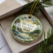 A decorative ornament in a box, featuring a scenic landscape with a waterfall, pine trees, and a cabin.