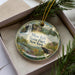 A decorative ornament with a scenic landscape of a waterfall, pine trees, and a cliff, placed inside a box with a pine branch.