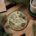 A decorative ornament featuring illustrations of Missouri's historic sites, including a mill, a log cabin, and a mining operation, is displayed on a wooden surface alongside a cup of coffee and some pine branches.