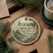 A decorative ornament with a scenic landscape of the Molalla River State Park in Oregon, featuring a waterfall and pine trees, is displayed on a wooden surface alongside a cup of coffee and some papers.