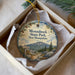 A decorative ornament in a box, featuring a scenic mountain landscape with pine trees and a trail sign.