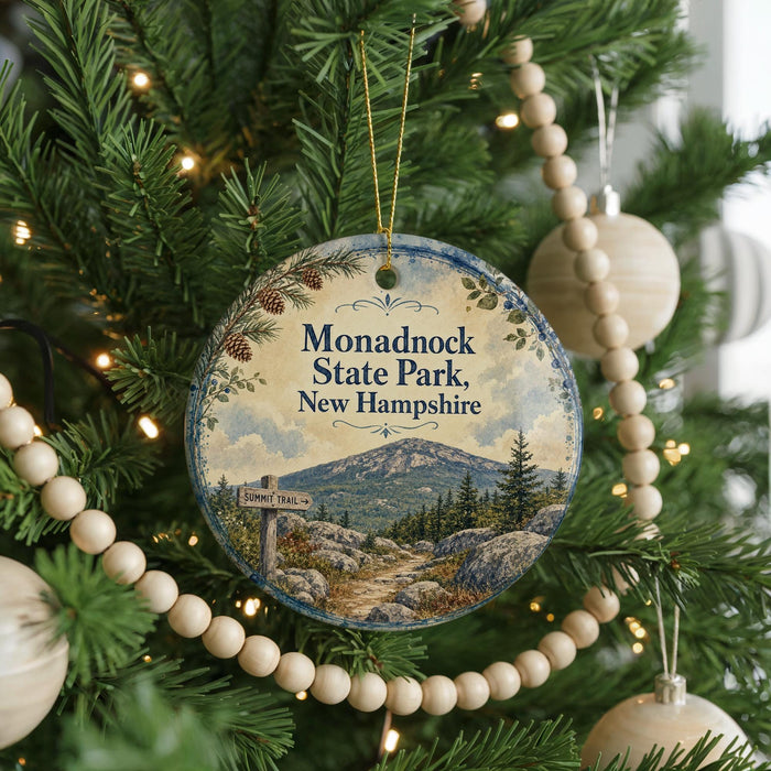 A round ornament hanging on a Christmas tree, featuring a scenic mountain landscape with pine trees and a trail sign.