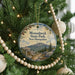 A round ornament hanging on a Christmas tree, featuring a scenic mountain landscape with pine trees and a trail sign.