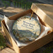 A decorative ornament in a box, featuring a scenic landscape with mountains and pine trees, with the text "Monadnock State Park New Hampshire" printed on it.