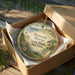 A decorative ornament in a box, featuring a lighthouse and a scenic landscape, with the text "Montauk Dows State Park, New York" printed on it.