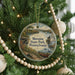A round ornament hanging on a Christmas tree, featuring a scenic landscape with a river, bridge, and boats.