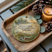 A decorative ornament with a scenic landscape of Mormon Island, the State Recreation Area in Nebraska, featuring a bird, pine cones, and a candle on a wooden tray.