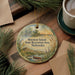 A round ornament with a scenic landscape depicting a lake, mountains, and a boat, hanging on a wooden surface.