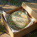 A round ornament in a box, featuring a scenic landscape with a lake, mountains, and a bird, is displayed on a wooden surface.