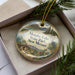 A decorative ornament in a box, featuring a scenic landscape with mountains, pine trees, and a lake.
