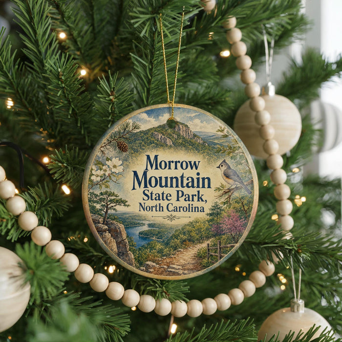 A round ornament hanging on a Christmas tree, featuring a scenic landscape with mountains, trees, and a winding path.