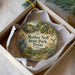 A decorative ornament in a box, featuring a scenic landscape with a house, trees, and wildlife, including a deer.