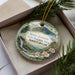 A decorative ornament featuring a scenic mountain landscape with a waterfall, surrounded by pine branches and pink flowers.