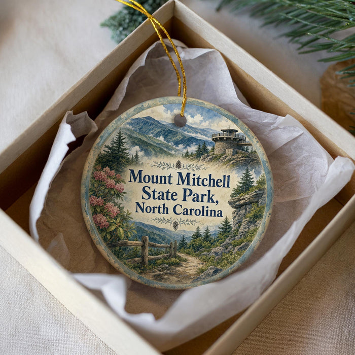 A decorative ornament in a box, featuring a scenic landscape with mountains, pine trees, and a castle-like structure.