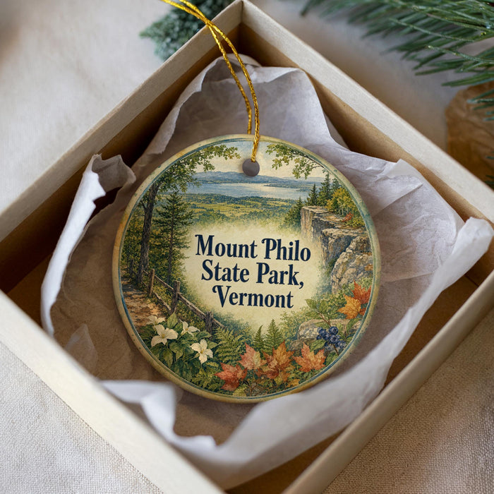 A decorative ornament in a box, featuring a scenic landscape with mountains, trees, and a path leading to a cliff.