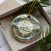 A decorative ornament in a box, featuring a scenic landscape with mountains, pine trees, and a waterfall.