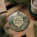 A decorative ornament featuring a scenic mountain landscape with pine trees, a waterfall, and a path, hanging on a wooden surface.