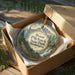 A round ornament in a box, featuring a scenic mountain landscape with a waterfall, surrounded by greenery.