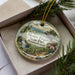 A decorative ornament in a box, featuring a scenic landscape with a waterfall and pine trees, with the text "Mount Pisgah State Park, Pennsylvania" printed on it.