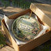 A round ornament in a box, featuring a scenic landscape with a waterfall and pine trees, with the text "Mount Pisgah State Park, Pennsylvania" displayed on it.