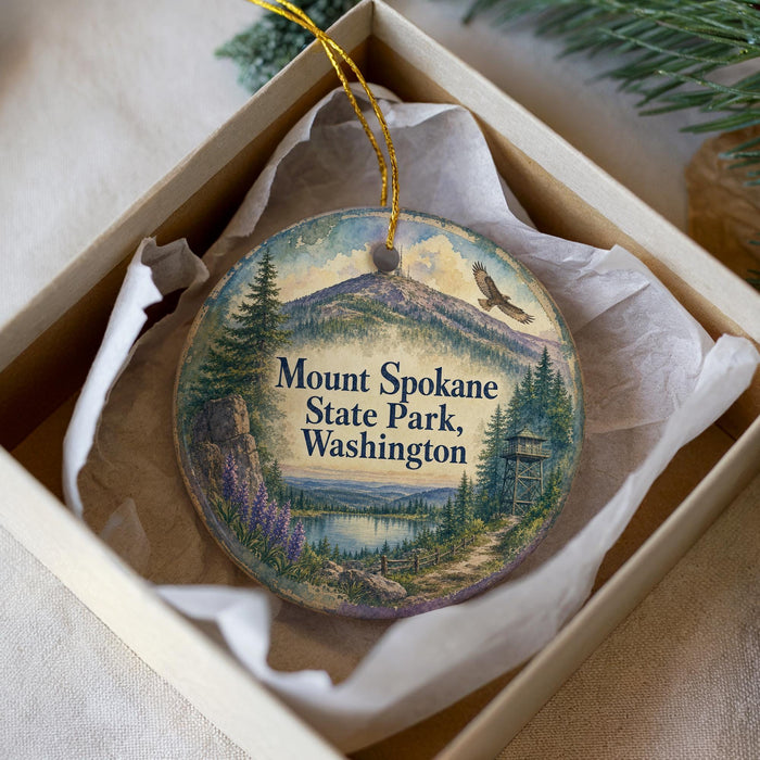 A decorative ornament in a box, featuring a scenic landscape with mountains, pine trees, and a lake.