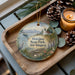 A decorative ornament featuring a scenic landscape with a lighthouse, mountains, and pine cones, placed on a wooden tray alongside a lit candle.