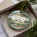 A decorative ornament in a box, featuring a beach scene with palm trees, a bird, and a body of water.