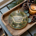 A decorative ornament with a scenic image of a park, featuring a path, trees, and wildlife, is displayed on a wooden tray alongside a lit candle and pine cones.