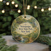 A round ornament with a painting of a river and park scenery, featuring a bird and a turtle, hangs in front of a blurred background with twinkling lights.