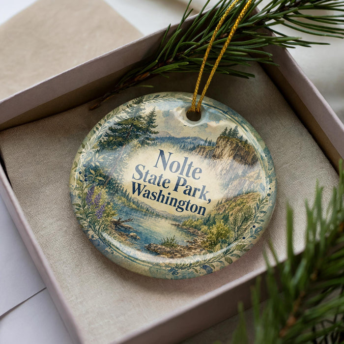 A decorative ornament in a box, featuring a scenic landscape with mountains, water, and pine trees.