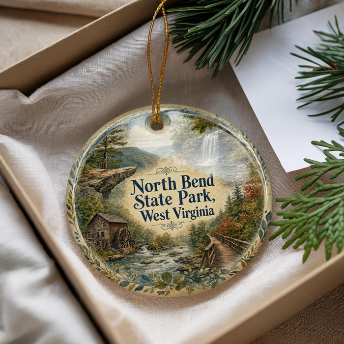 A decorative ornament with a scenic landscape of North Bend State Park, West Virginia, featuring a river, waterfall, and a wooden cabin.