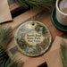 A decorative ornament featuring a scenic image of North Bend State Park, West Virginia, with a waterfall and a cabin in the background. The ornament is hanging on a wooden surface, surrounded by pine branches and a cup of coffee.