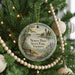 A round ornament hanging on a Christmas tree, featuring a scenic landscape with a lighthouse and a bench.