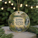 A round ornament with a painting of a scenic landscape, featuring a waterfall, trees, and a bird, hangs in front of a Christmas tree with lights.