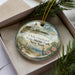 A decorative ornament with a lighthouse and a coastal scene, placed in a box with pine branches.
