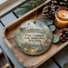 A decorative ornament featuring a lighthouse and a serene coastal scene, placed on a wooden tray alongside pine cones and a lit candle.