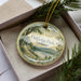 A decorative ornament in a box, featuring a scenic landscape with a lake, mountains, and a park.