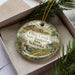 A decorative ornament in a box, featuring a scenic landscape with a mountain, river, and trees.