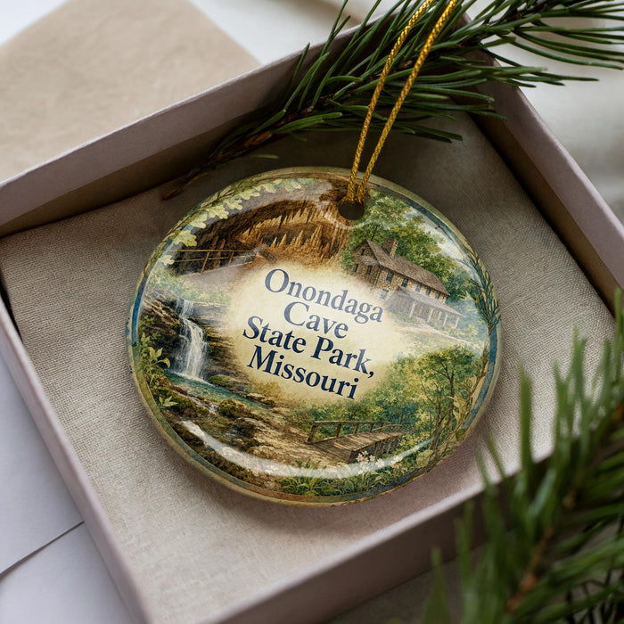 A decorative ornament with a scenic image of the Onondaga Cave in Missouri, featuring a waterfall and a cabin, is displayed in a box with pine branches.