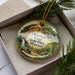 A decorative ornament with a scenic image of the Onondaga Cave in Missouri, featuring a waterfall and a cabin, is displayed in a box with pine branches.