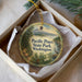 A decorative ornament in a white box, featuring a scenic landscape with a mountain, pine trees, and a body of water.