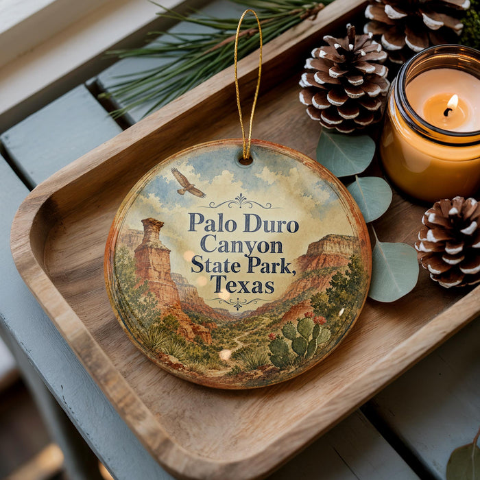 A decorative ornament with a scenic image of the Palo Duro Canyon State Park in Texas, featuring a bird and desert landscape, is displayed on a wooden tray alongside pine cones and a lit candle.