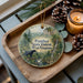 A decorative ornament featuring a black panther in a forest setting, with the text "Panther State Forest, West Virginia" printed on it. The ornament is placed on a wooden tray, surrounded by pine cones and a lit candle.