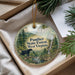A decorative ornament with a black bear standing on a rock, surrounded by a forest scene, is displayed in a box with pine branches.