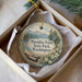 A decorative ornament in a white box, featuring a scenic landscape with mountains, pine trees, and a body of water.