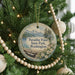 A round ornament hanging on a Christmas tree, featuring a scenic landscape of mountains, pine trees, and a lake.