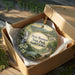 A decorative ornament in a box, featuring a scenic landscape with a waterfall and a cabin, with the text "Paris Mountain State Park, South Carolina" prominently displayed.