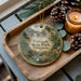 A decorative ornament featuring a scenic image of Patison State Park, Wisconsin, with a candle and pine cones on a wooden tray.