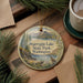 A decorative ornament featuring a scenic landscape of Pearyrgin Lake State Park, Washington, with a bird perched on a rock. The ornament is placed on a wooden surface, surrounded by pine branches and a cup of coffee.